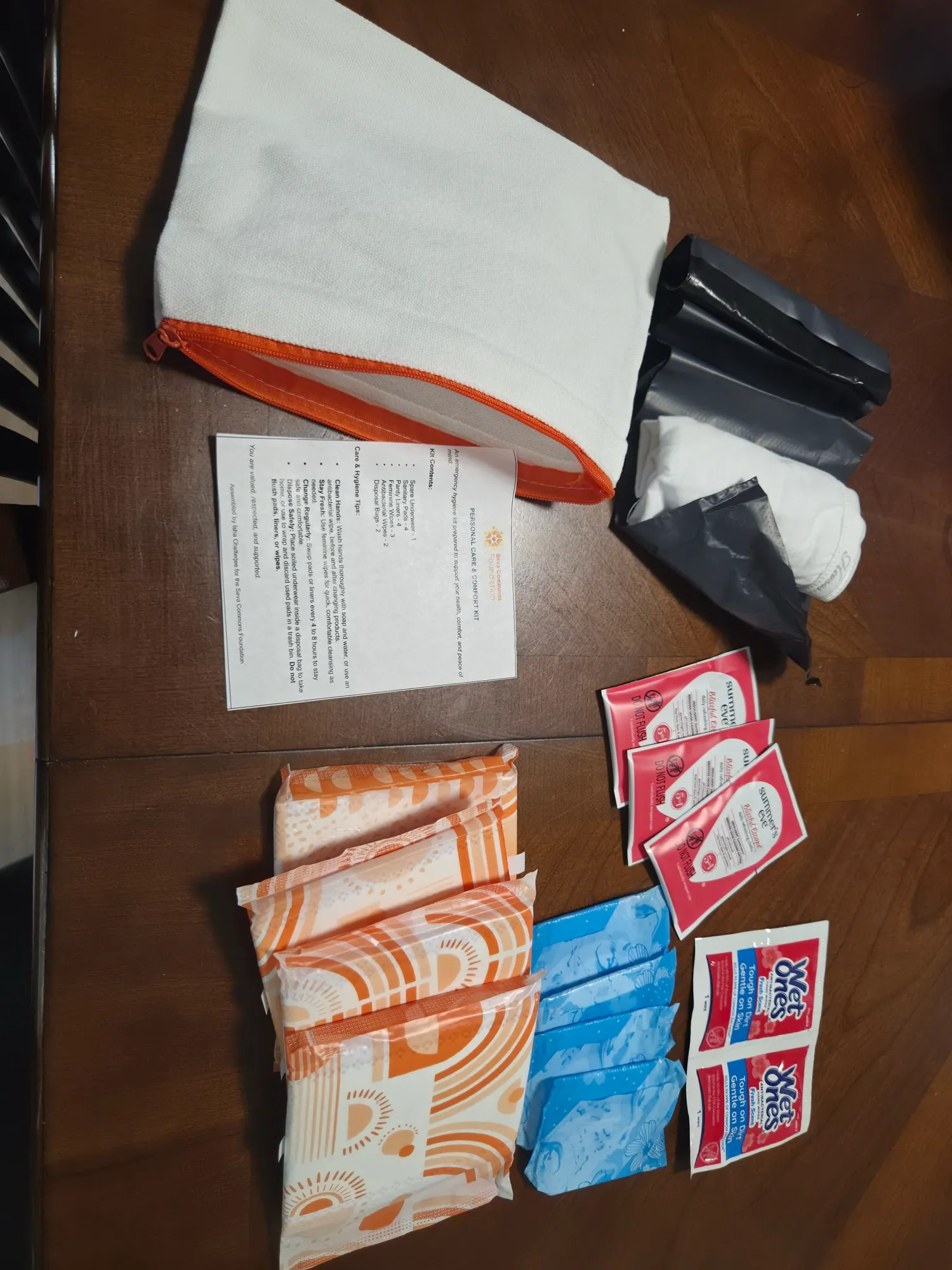 The contents of one hygiene kit laid out: a zippered canvas pouch, pads, wipes, opaque disposal bags, new cotton underwear, and a printed care card.