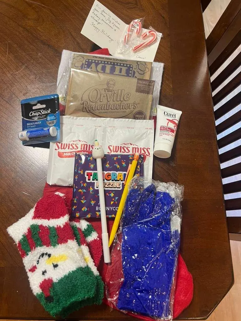 The contents of one stocking laid out: fuzzy holiday socks, gloves, hot chocolate, popcorn, candy canes, lip balm, lotion, a tangram puzzle, a pen and pencil, and a handwritten card.